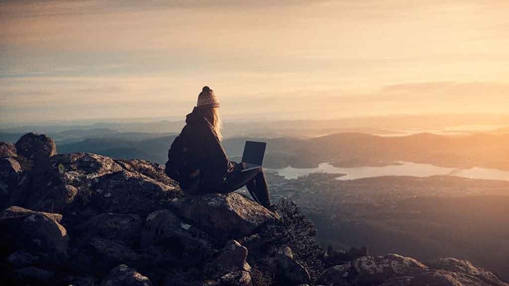 A woman with a laptop sits on a rock overlooking a valley, symbolizing being on the cusp of tapping into the potential of AI in supply chains and AI.