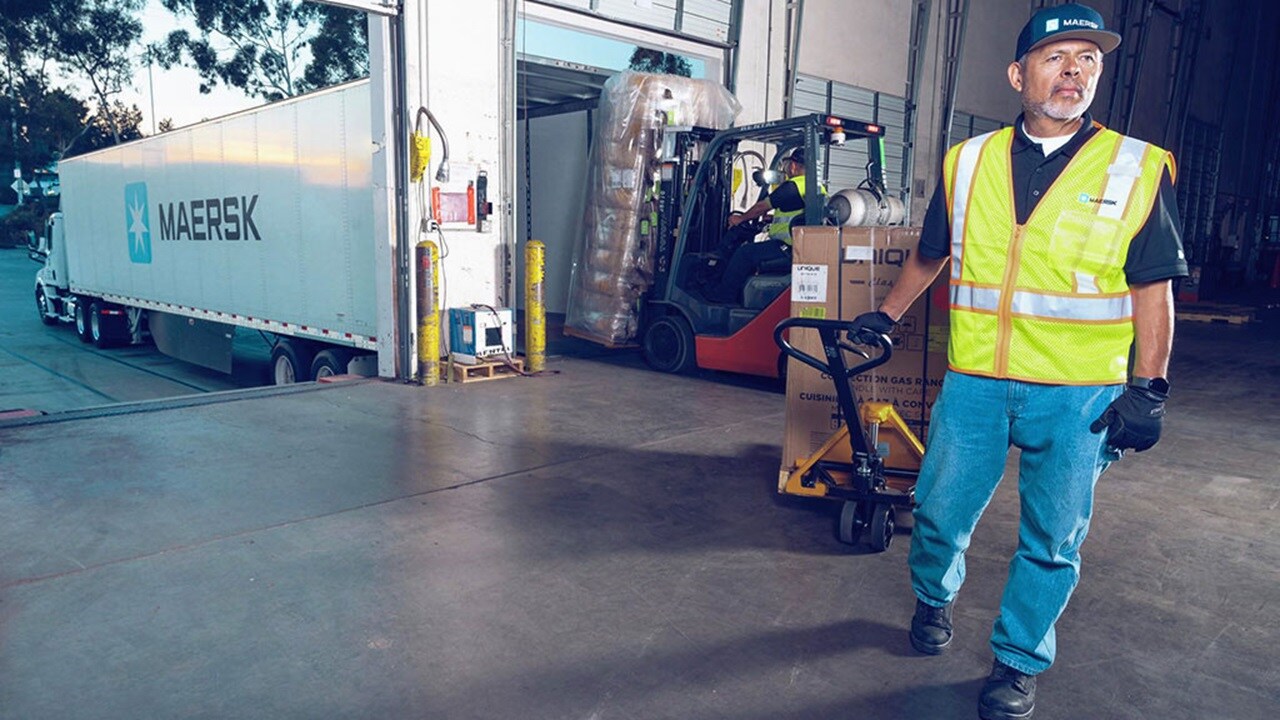 Person in a high-visibility vest using a manual pallet jack to move a pallet loaded with boxes