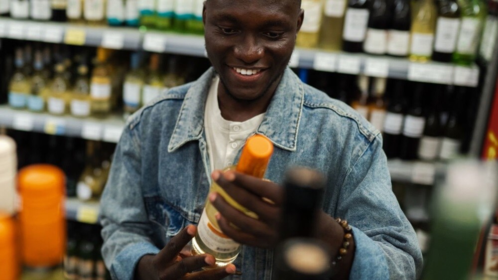 Smiling customer in denim jacket choosing a wine bottle in liquor store aisle.
