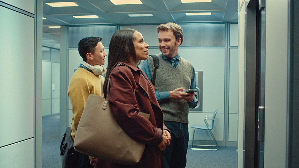 A group of three people stands inside an elevator, with modern office decor in the background. Two individuals appear engaged in conversation.