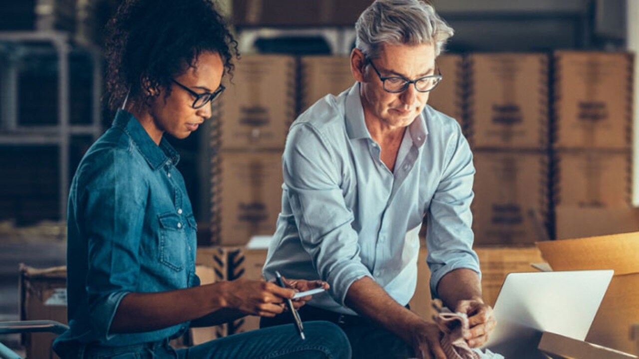 A couple of employees discussing in a warehouse with small boxes in the background.