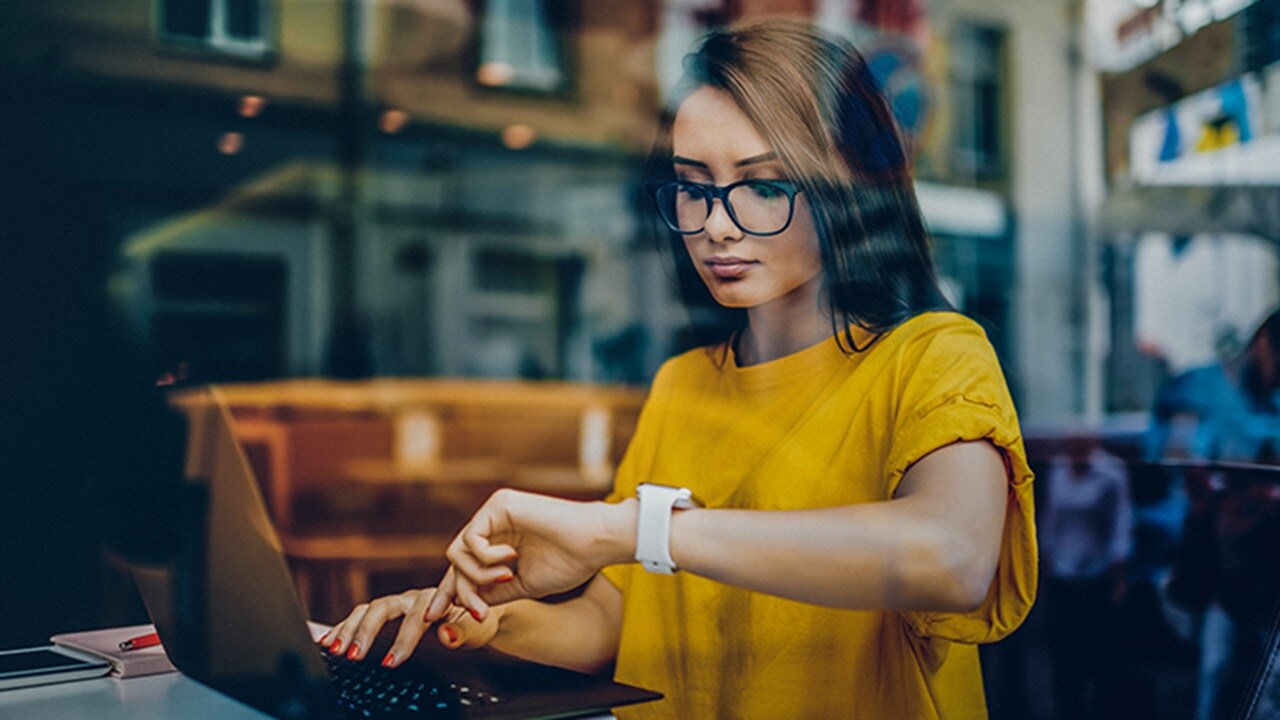 A woman in a warehouse typing on a laptop with boxes and pallets in the background.